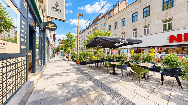 Lebhafte Straße mit Geschäften und einem Außenbereich mit Tischen und Stühlen unter einem Sonnenschirm.