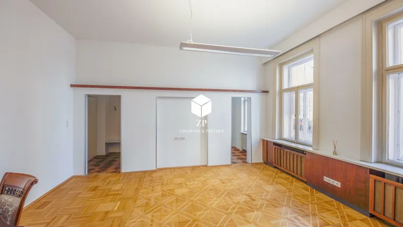 Geräumiger Raum mit Fischgrätparkett, hellen Wänden und großen Fenstern mit Holzverkleidung, ideal für Wohn- oder Bürozwecke.