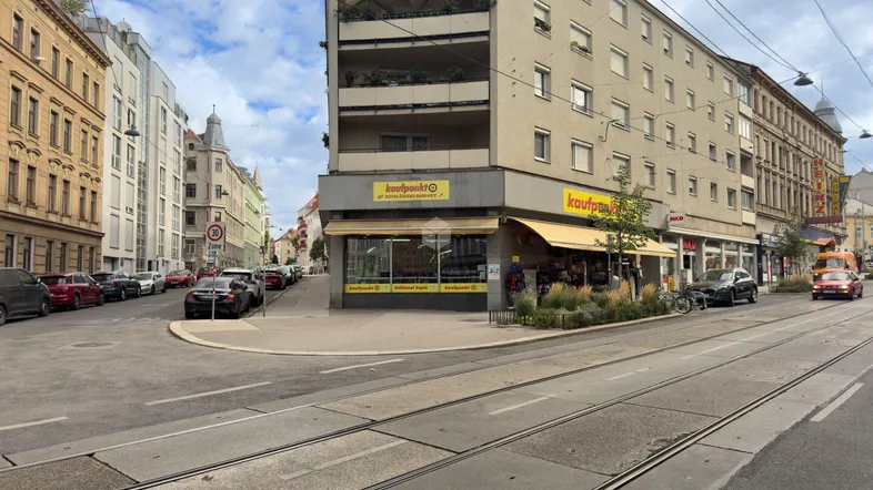 Eine belebte Straßenszene mit einem Eckgebäude, Geschäften im Erdgeschoss und Straßenbahnschienen im Vordergrund.