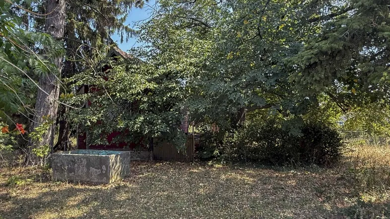 Kleines rotes Gebäude umgeben von Bäumen und hohem Gras in ländlicher Umgebung.