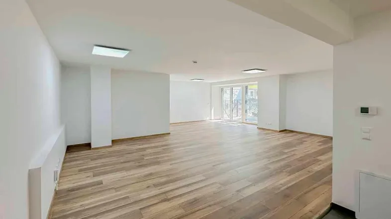 Geräumiger, leerer Raum mit hellem Holzboden und großen Glastüren, die viel Tageslicht hereinlassen.