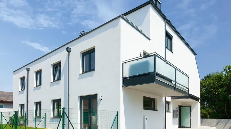 Modernes Mehrfamilienhaus mit weißer Fassade, Balkon und gepflegter Außenanlage unter blauem Himmel.