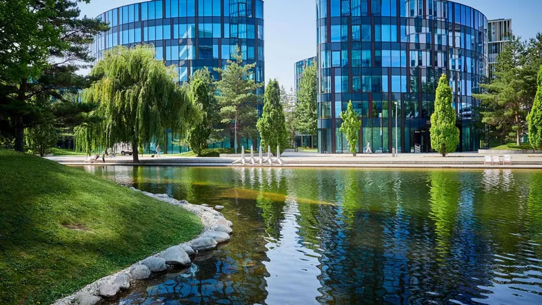 Moderne Bürogebäude mit blauer Glasfassade und einem Teich in einer grünen Parklandschaft unter blauem Himmel.
