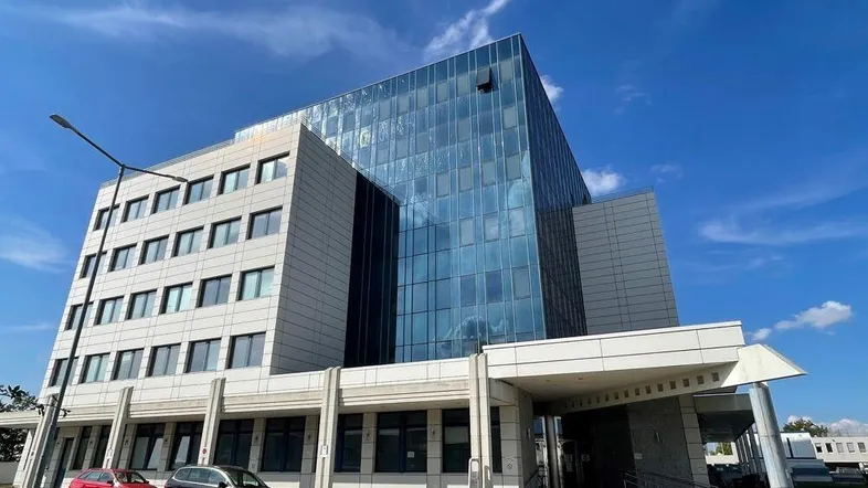 Modernes Bürogebäude mit Glasfassade und hellen Steinplatten unter blauem Himmel und leichter Bewölkung.