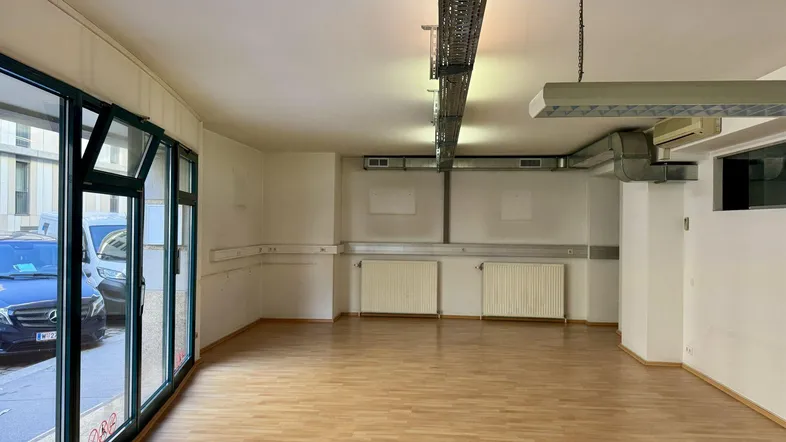Heller Gewerberaum mit großen Schaufenstern, Laminatboden und sichtbarer Deckeninstallation.