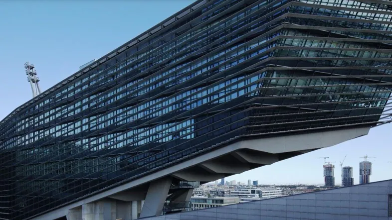 Moderne, markante Architektur mit großer Glasfassade und städtischem Hintergrund unter blauem Himmel.