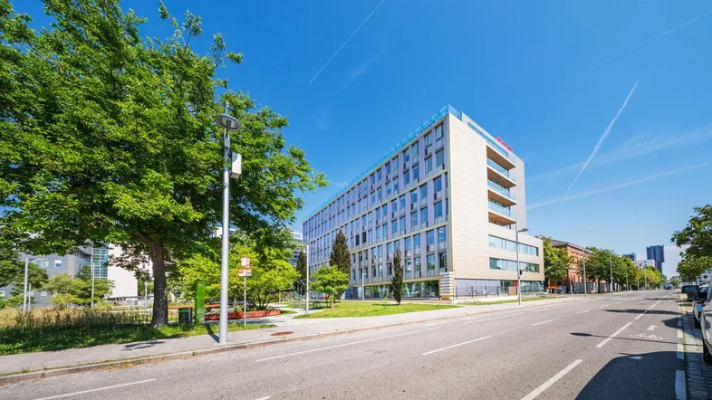 Modernes Bürogebäude mit vielen Fenstern und Balkonen an einer belebten Straße unter blauem Himmel.