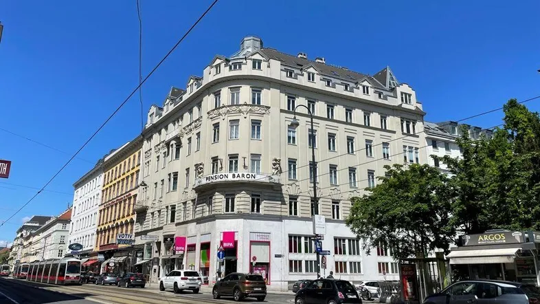 Historisches Gebäude an einer belebten Straße mit Straßenbahnen und Geschäften unter blauem Himmel.