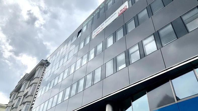 Modernes Bürogebäude mit dunkler Fassade und vielen Fenstern unter bewölktem Himmel.