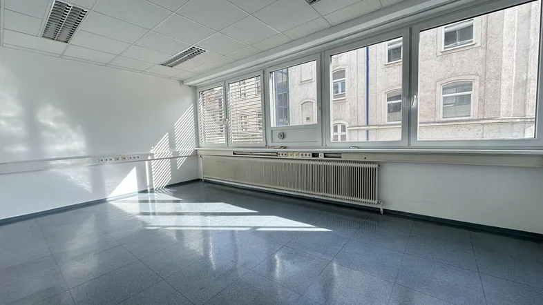 Helles, leeres Büro mit großen Fenstern, die viel Tageslicht hereinlassen und Blick auf andere Gebäude bieten.