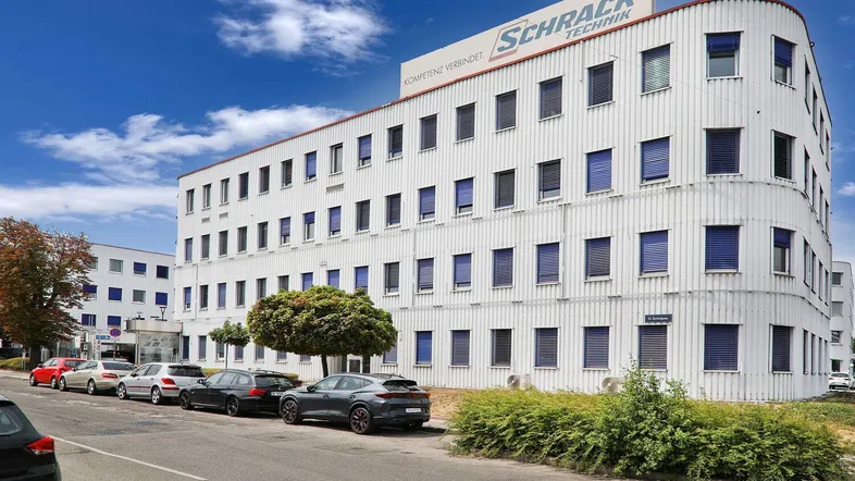Großes, modernes Bürogebäude mit weißer Fassade und blauen Fensterläden, umgeben von Parkplätzen und Bäumen.