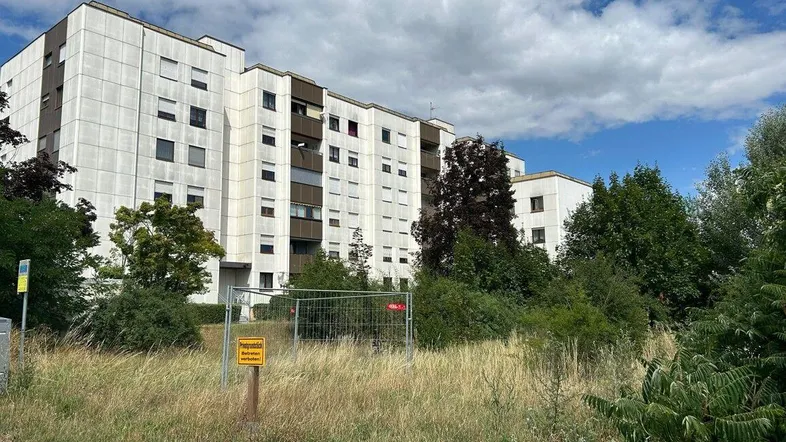 Mehrstöckiges Wohngebäude mit Balkonen im Hintergrund, umgeben von einer Wiese und Bäumen unter blauem Himmel.