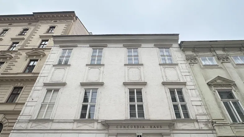 Historische Fassade des Concordia-Hauses in einer Reihe von Gebäuden unter bewölktem Himmel.