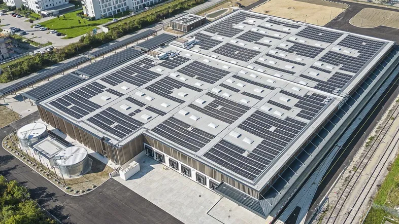 Luftaufnahme eines großen Gewerbegebäudes mit umfangreicher Photovoltaikanlage auf dem Dach und umliegender Infrastruktur.