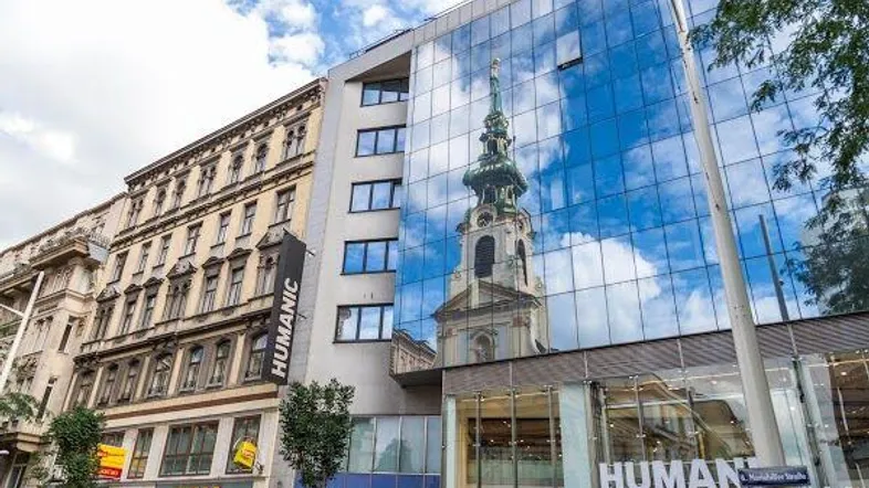 Moderne Glasfassade eines Geschäftsgebäudes, die einen historischen Kirchturm und ein angrenzendes Altbaugebäude widerspiegelt.