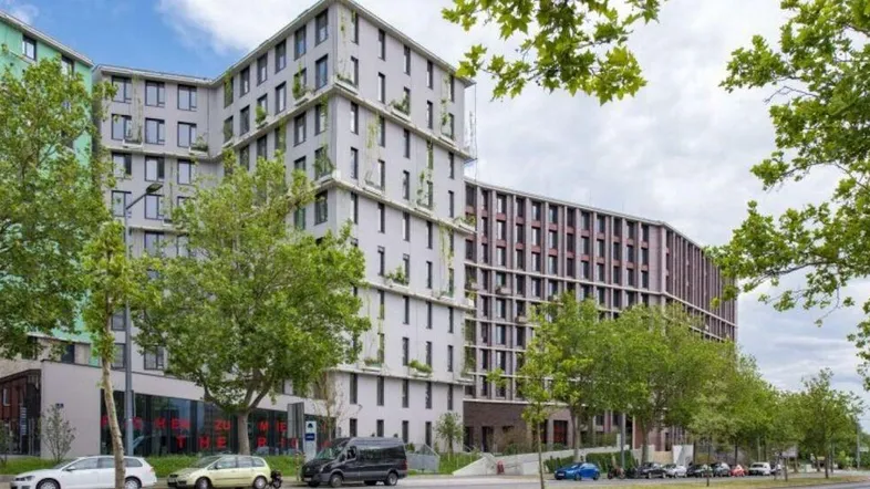 Modernes Gebäude mit vielen Fenstern und begrünten Fassadenelementen, umgeben von Bäumen und einer Straße.
