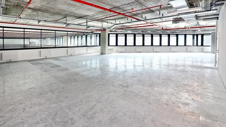 Großer, offener Büroraum mit unverputzter Betondecke, sichtbaren Leitungen und großen Fenstern, bereit für den Ausbau.