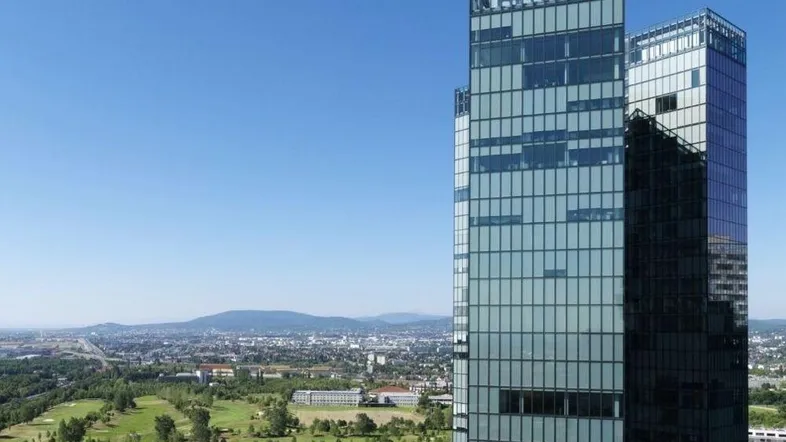 Moderne Wolkenkratzer mit Glasfassaden ragen über einer grünen Stadtlandschaft mit Bergen im Hintergrund auf.