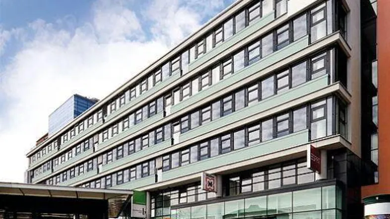 Modernes Geschäftsgebäude mit vielen Fenstern und Glasfassade im Erdgeschoss, belebtem Gehweg davor.