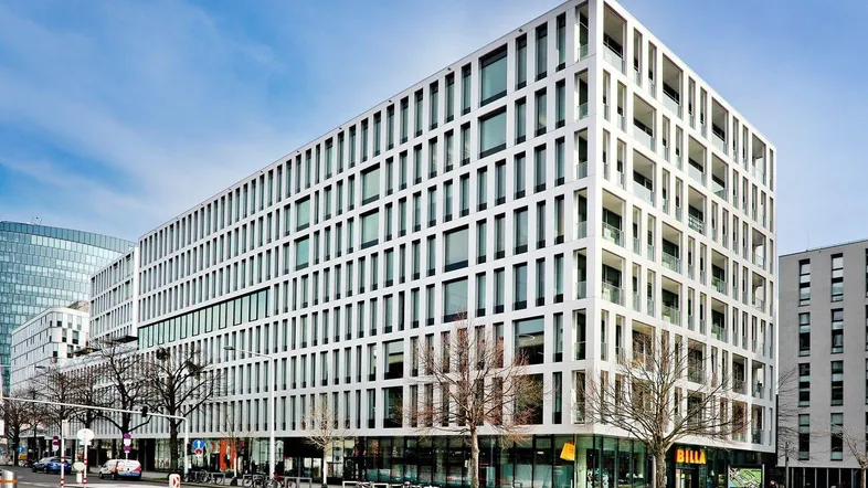 Modernes Bürogebäude mit markanter weißer Fassade und großen Fenstern an einer städtischen Straße. Im Erdgeschoss befinden sich Einzelhandelsflächen.
