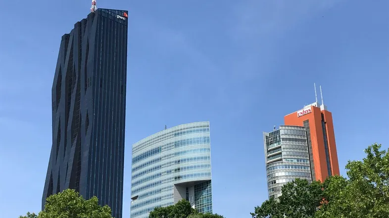 Moderne Hochhäuser mit markanter Architektur und Glasfassaden vor blauem Himmel, umgeben von grünen Bäumen.