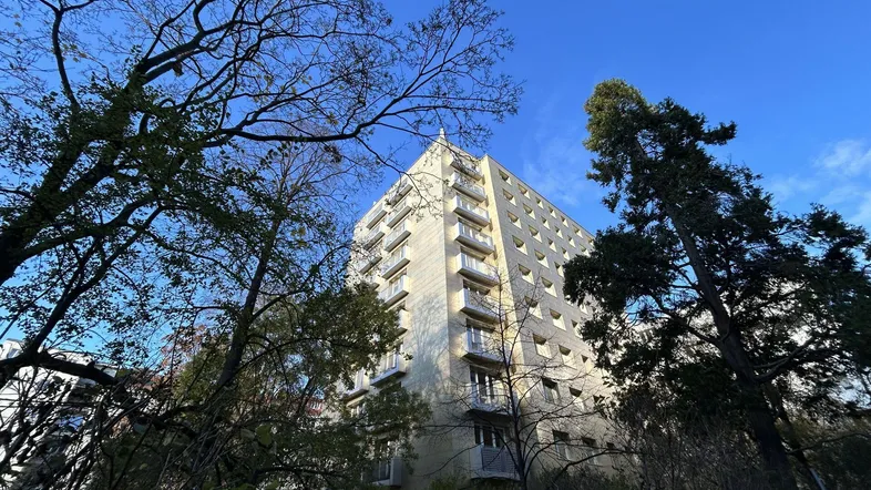 Hohes Wohngebäude mit mehreren Stockwerken und Balkonen, umgeben von Bäumen unter blauem Himmel.
