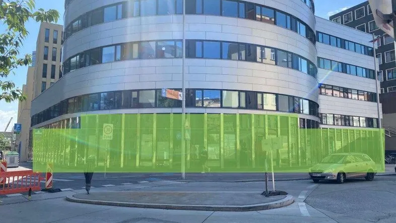 Modernes Bürogebäude mit runder Fassade und großen Fenstern an einer städtischen Straße, teilweise von einer grünen Barriere verdeckt.