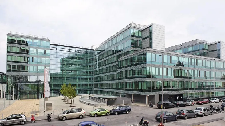 Modernes Bürogebäude mit Glasfassade und Parkplätzen davor unter bewölktem Himmel.