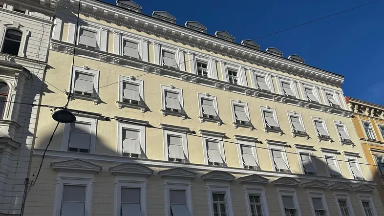 Fassade eines historischen Gebäudes mit vielen Fenstern und Jalousien unter blauem Himmel.