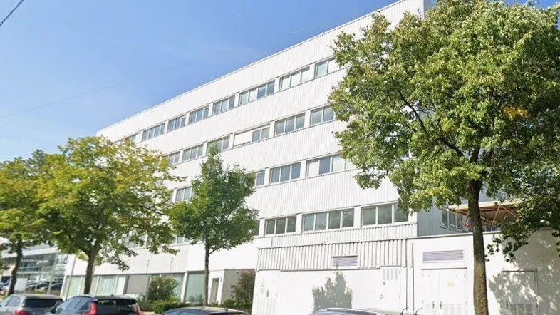 Modernes Bürogebäude mit weißer Fassade und zahlreichen Fenstern, umgeben von Bäumen und parkenden Autos.