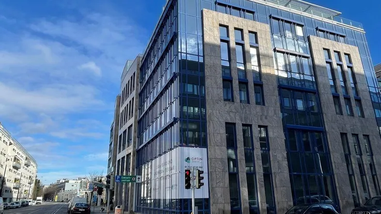 Modernes Bürogebäude mit Glasfassade und Steinelementen an einer belebten Straße unter blauem Himmel.