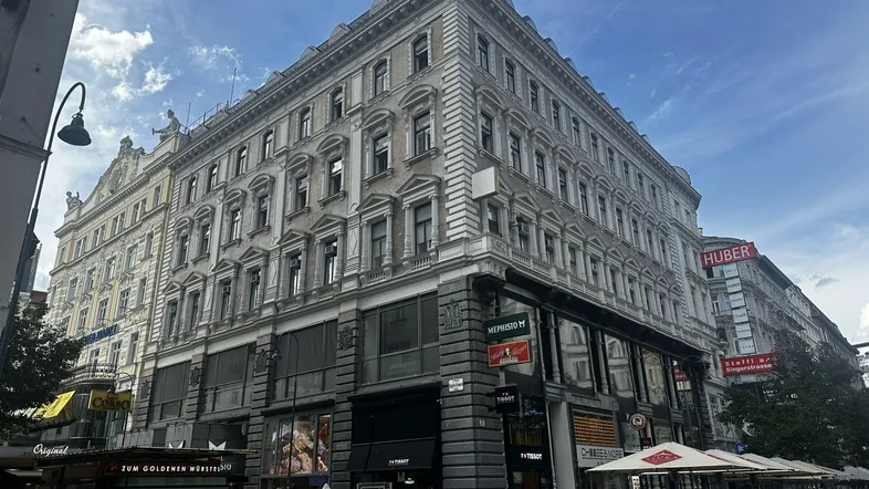Historisches Gebäude mit reich verzierter Fassade an einer belebten Stadtstraße unter bewölktem Himmel.