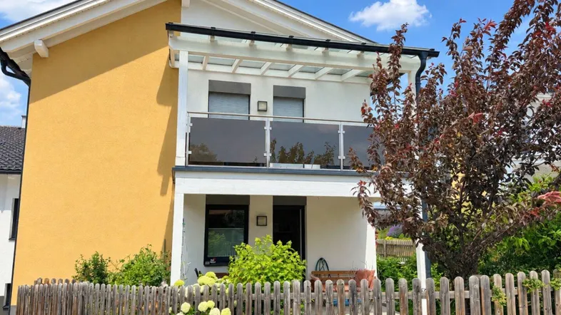 Modernes Wohnhaus mit gelber Fassade, Balkon mit Glasgeländer und kleinem Vorgarten.