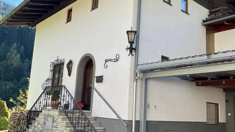 Traditionelles Haus mit weißer Fassade, Steintreppe und Carport in alpiner Umgebung.