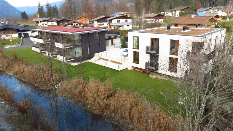 Moderne Gebäude in alpiner Umgebung mit Flusslauf und Bergkulisse im Hintergrund.