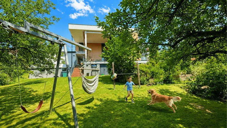 Sonnige Rasenfläche im Garten mit spielendem Kind und Hund vor dem Haus.