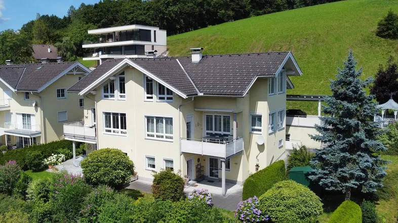 Luftaufnahme eines hellgelben Hauses mit Satteldach, umgeben von grünen Hügeln und Bäumen.