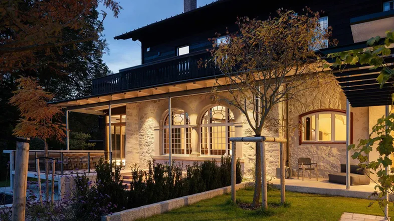Elegante Villa mit Steinfassade, überdachter Terrasse und gepflegtem Gartenbereich am Abend.