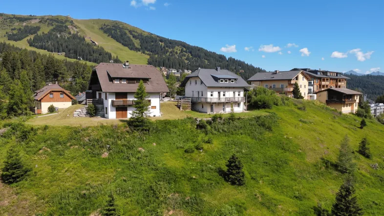 Mehrere Häuser in einer malerischen Berglandschaft mit grünen Hängen und Wäldern unter blauem Himmel.