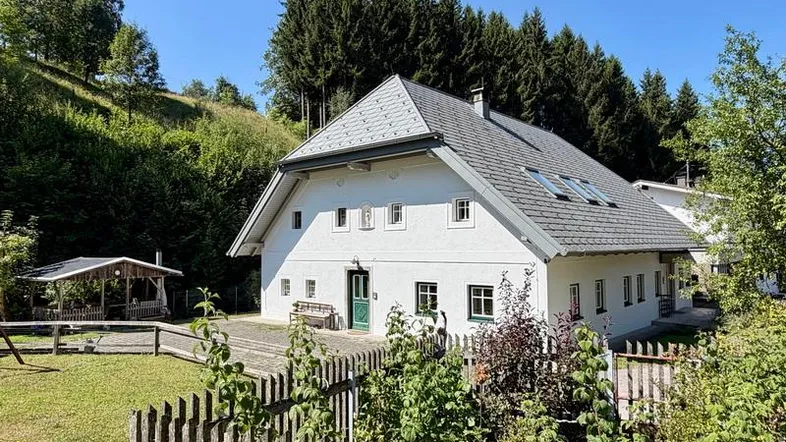 Weißes Landhaus mit Satteldach, umgeben von grünen Hügeln und Wald, mit einem Holzzaun im Vordergrund.