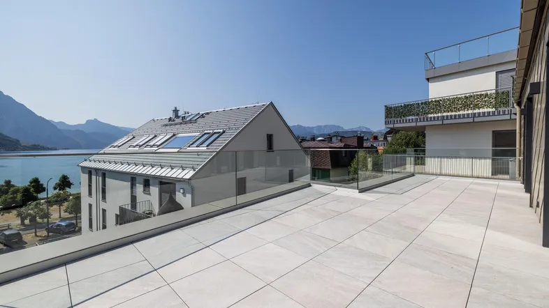 Große Dachterrasse mit Fliesenboden, Glasgeländer und weitem Blick auf den See und die Berge.