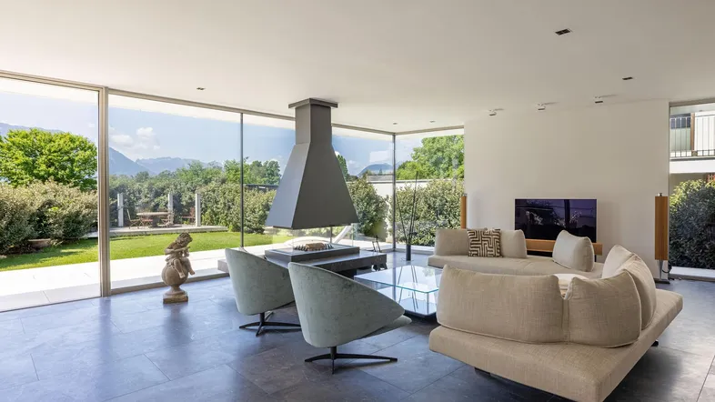 Helles, modernes Wohnzimmer mit Kamin, großen Glasfronten und Blick auf den Garten und die Berge.