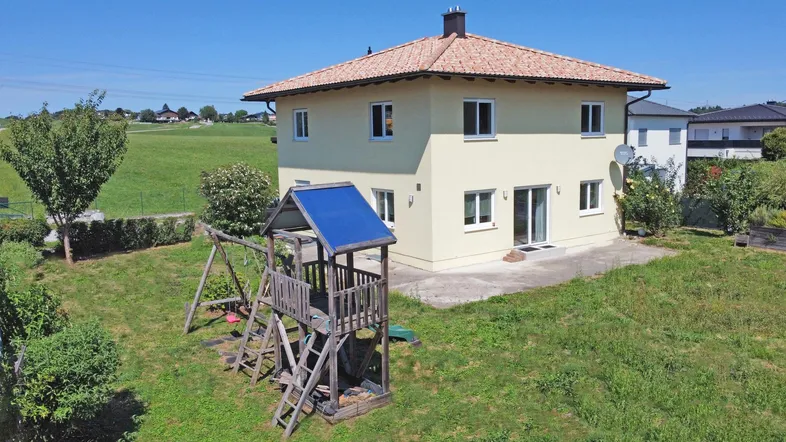 Einfamilienhaus mit großem Garten und Kinderspielplatz unter blauem Himmel.