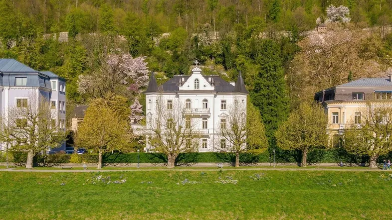 Majestätische weiße Villa am Flussufer mit üppiger grüner Hügellandschaft im Hintergrund.
