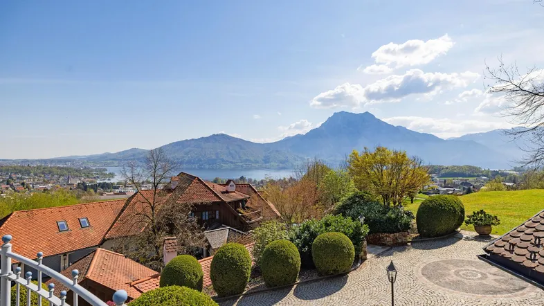 Panoramablick von einem Balkon auf einen See, Berge und die umliegende Landschaft unter blauem Himmel.