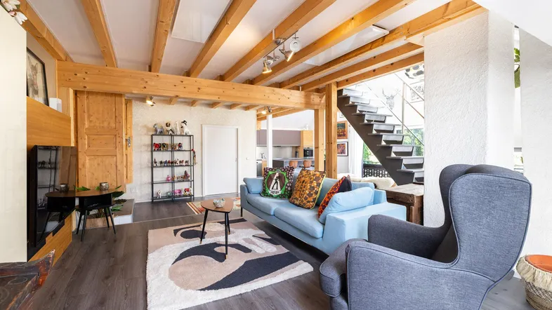 Helles Wohnzimmer mit sichtbaren Holzbalken, modernem Sofa und offenem Treppenaufgang.