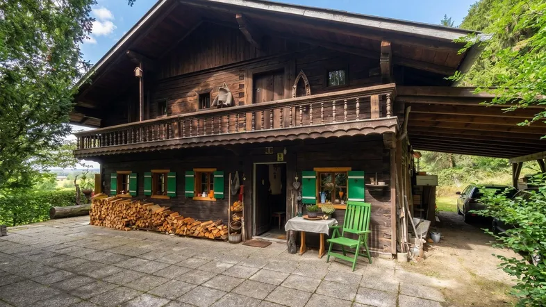 Traditionelles Holzchalet mit Balkon, Holzstapel und gepflastertem Vorplatz in ländlicher Umgebung.