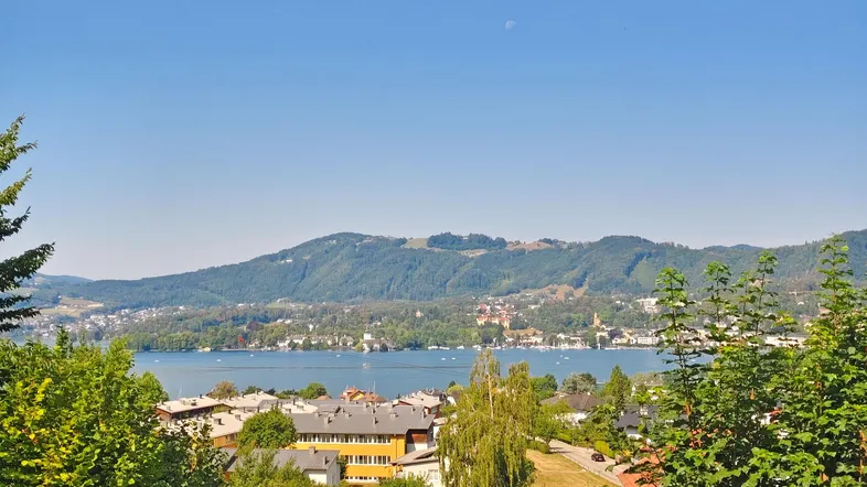 Panoramablick auf den See und die umliegenden Berge mit Häusern am Ufer unter blauem Himmel.