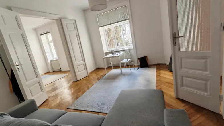 Helles Wohnzimmer mit Fischgrätparkett, einem grauen Sofa und einem Arbeitsbereich am Fenster.