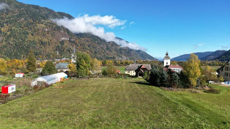 Luftaufnahme eines weitläufigen grünen Feldes mit herbstlichen Bäumen und Bergen im Hintergrund.